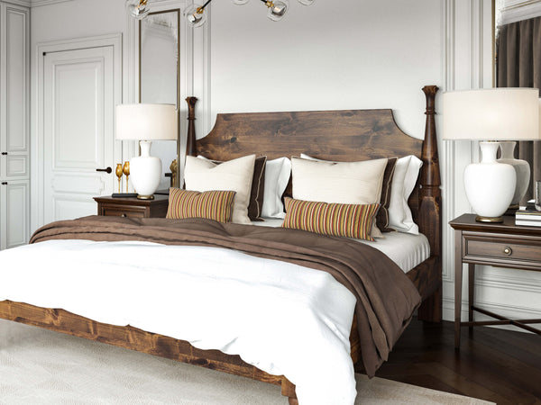 A cozy bedroom with a dark wooden bed frame, cream and beige pillows, and a plaid blanket. The room is softly lit with a table lamp and has sunlight streaming through wooden blinds on the window.