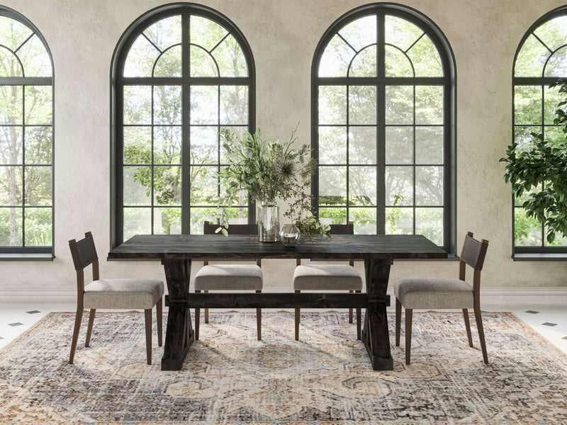 A dining room features the Trestle Dining Table - Charred Ember by James+James, accompanied by four beige upholstered chairs on a patterned rug. A bouquet of greenery in a glass vase adorns the center. Large arched windows with black frames provide abundant natural light, showcasing a lush green outdoor scene.
