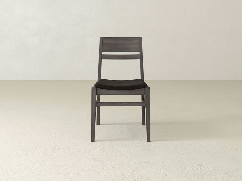 The Delilah Dining Chair - Deep Grey from James+James, a straightforward and contemporary wooden chair with a dark finish and a black cushioned seat, is placed against a plain light beige background. The chair showcases a horizontal slat back design and clean, minimalist lines.