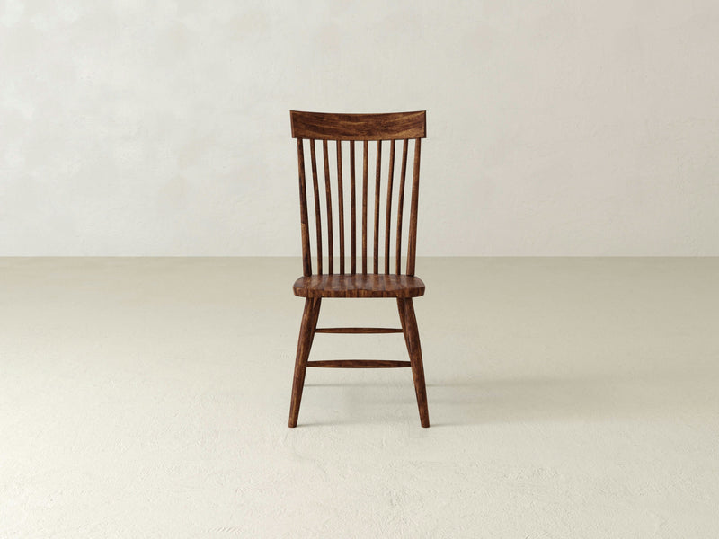 An Eleanor Side Dining Chair - Tuscany by James+James, featuring a wooden slatted backrest and minimalistic design, stands on a smooth, neutral-colored floor against a light gray wall. The chair showcases a natural wood finish that highlights its grain.