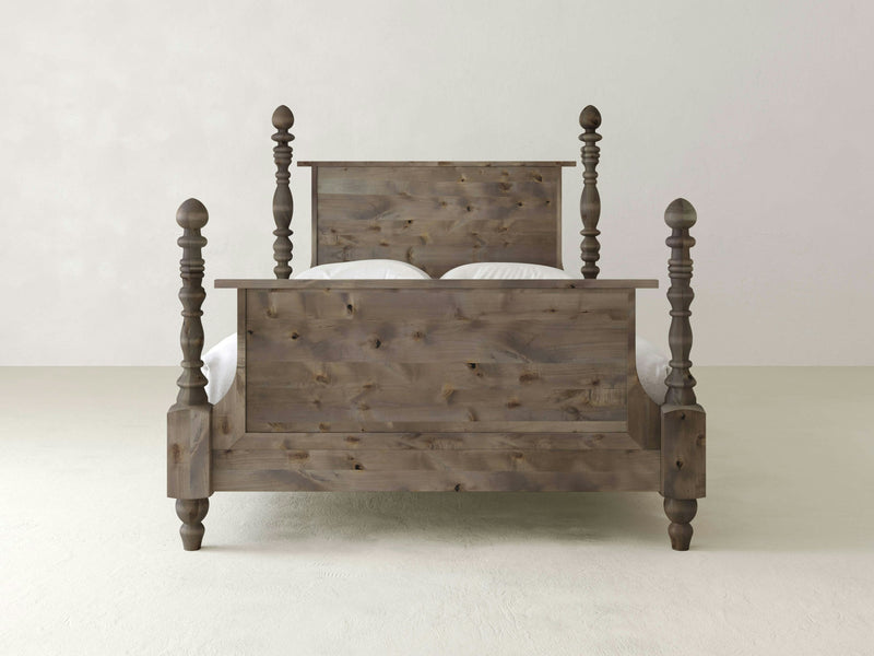 The Catherine Bed - Barn Wood by James+James is a rustic wooden bed featuring a headboard and footboard with four turned posts at each corner. It has a natural, distressed finish that lends it a vintage or farmhouse aesthetic. The minimalist background consists of a white wall and a pale floor.