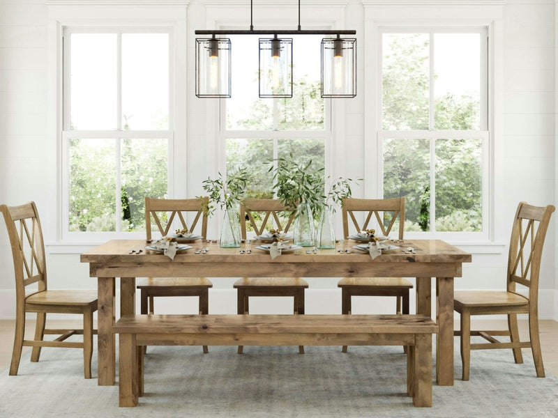 Bright dining room featuring a wooden table with a matching James+James Farmhouse Bench - Harvest Wheat and four chairs. The table is set with tableware and green plants as a centerpiece. A modern chandelier with four lights hangs above the table. Large windows in the background brighten the space.