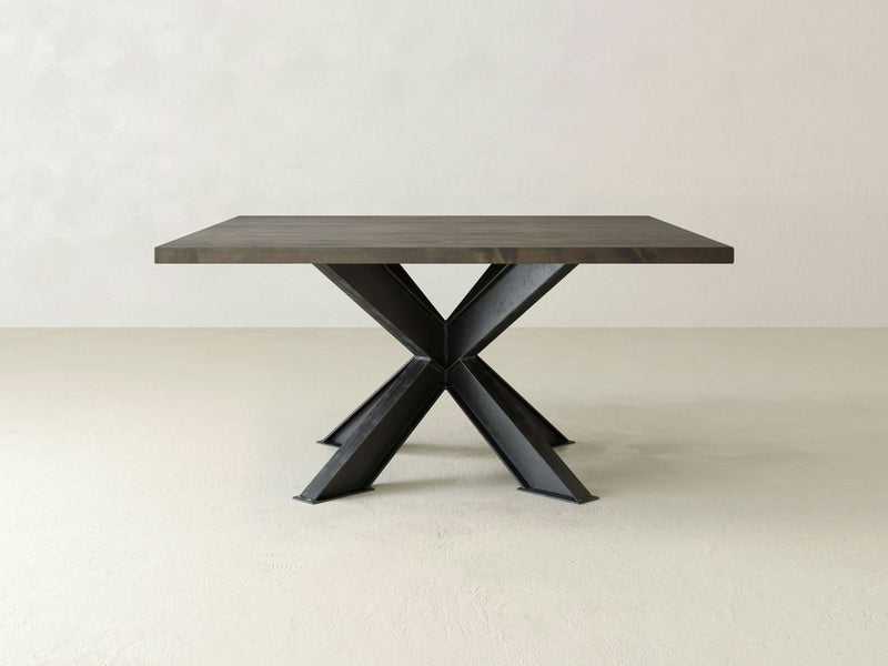 A modern square Shiloh Dining Table in deep grey finish from James+James. This wooden table features a thick tabletop and stands on a unique metal base with four angled legs that converge in the middle to form a striking X shape. The background is an off-white wall and floor.