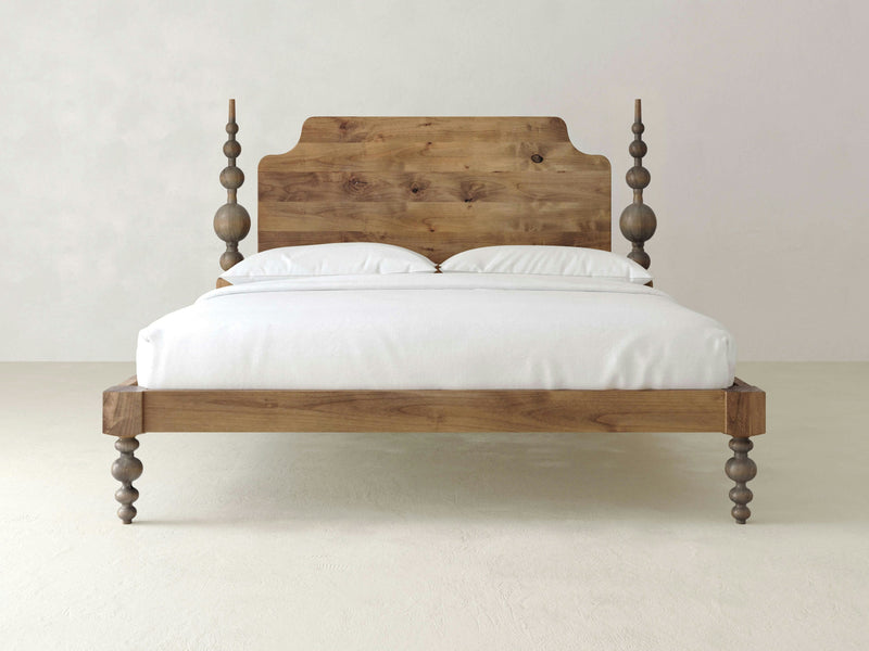 The Opal Bed - Harvest Wheat by James+James is a wooden bed featuring a high headboard with intricate spindle designs on each side. It comes with a white mattress and two white pillows, and stands on four turned wooden legs adorned with matching spindles. The background showcases a plain, light-colored wall and floor.