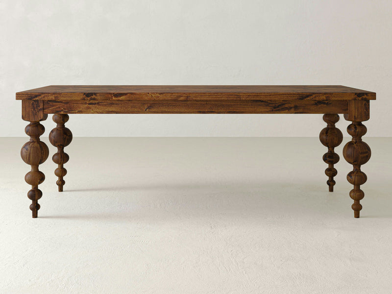 The Olivia Dining Table - Tuscany by James+James features a polished rectangular wooden surface and intricately carved legs. The table legs showcase a series of rounded, bulbous shapes stacked vertically, contributing to its decorative and ornate appearance. This elegant table stands alone in a minimalist setting.