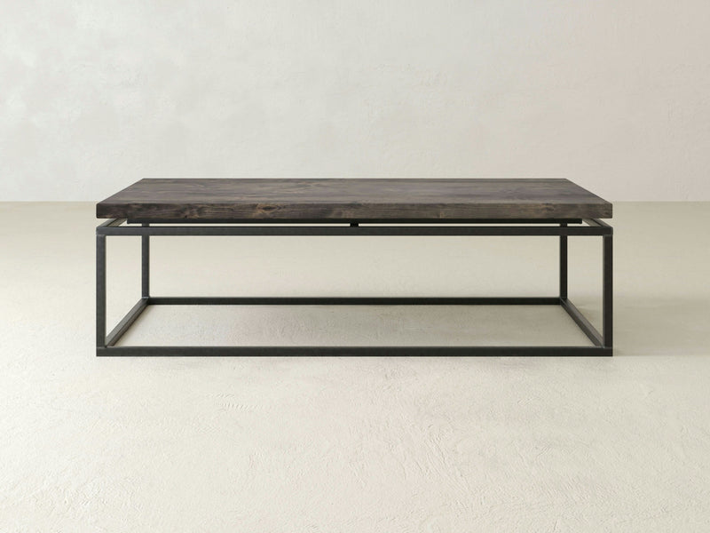 A minimalist James+James Floating Top Coffee Table - Deep Grey, featuring a rectangular wooden top and a sleek black metal open-frame base, is set against a plain white background. The design is clean and modern, emphasizing simplicity and functionality.