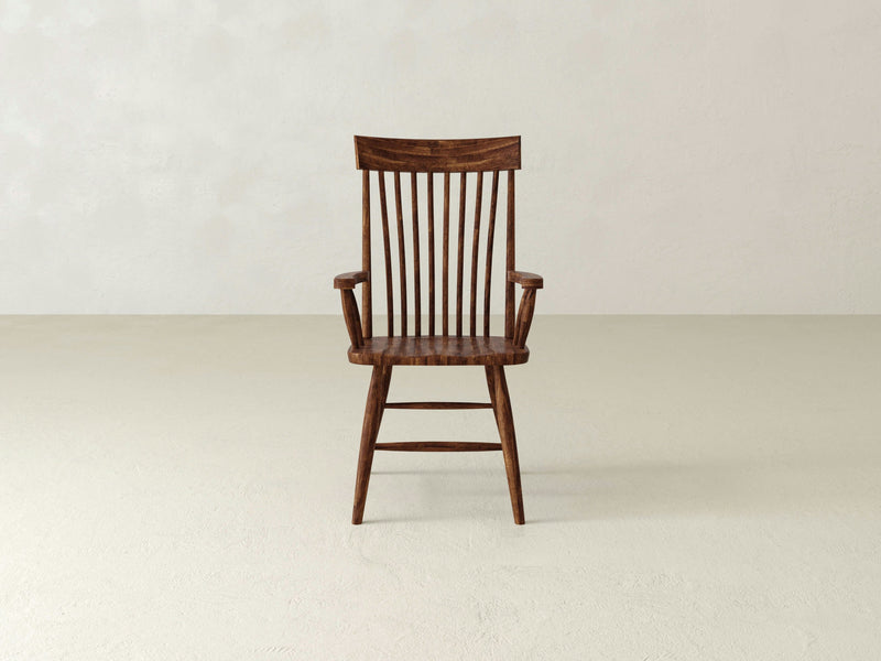 The Eleanor Arm Dining Chair - Tuscany by James+James, featuring a high backrest with vertical slats and armrests, is placed on a light-colored floor against a plain, off-white background. The wooden chair showcases a rustic finish with slightly tapered legs.