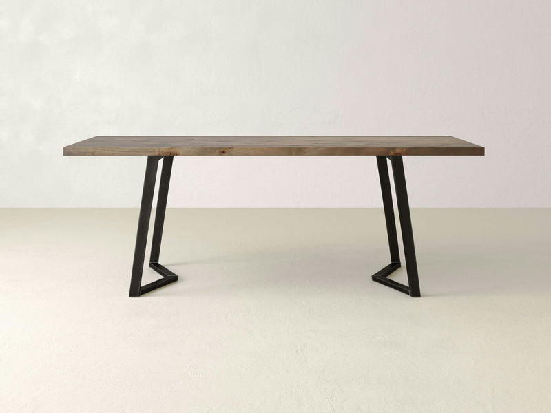 The Arkwright Dining Table - Barn Wood from James+James features a rectangular wooden top and sleek, black metal legs arranged in a V-shape on either end, set against a plain, light-colored background. This table embodies a minimalist and contemporary design.