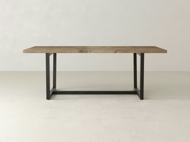 The Trapezoid Dining Table by James+James boasts a contemporary design with its rectangular wooden top featuring a natural finish and black metal legs. This table exudes an industrial chic appeal and is set against a plain, light-colored background.