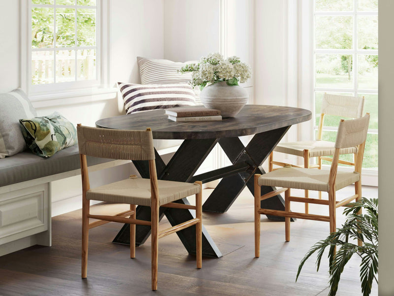 A sunlit dining area with a James+James X-Base Oval Dining Table in Tobacco. Surrounding the table are three chairs with light wooden frames and woven seats. A cushioned bench with pillows sits by the window. A large, leafy plant is in the corner.