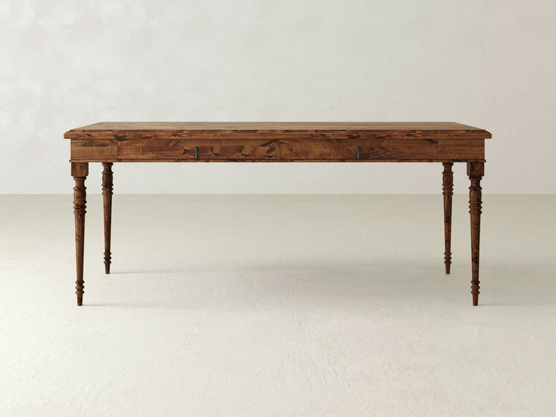 The "Audrey Desk - Tuscany" by James+James is a rectangular wooden desk featuring ornate, turned legs. The desk boasts a natural wood finish that beautifully showcases the grain and texture. It stands against a plain, light-colored floor and is set against a muted, light-colored wall background.