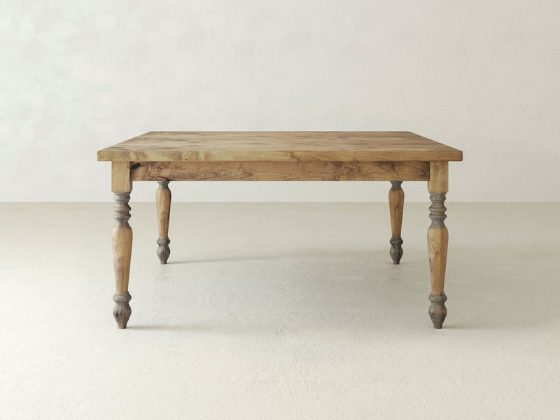 A French Country Square Dining Table - Harvest Wheat by James+James, featuring a natural finish, is positioned centrally against a plain, light-colored background. The table has a square top with slightly rounded edges and four turned legs with a decorative, carved design.