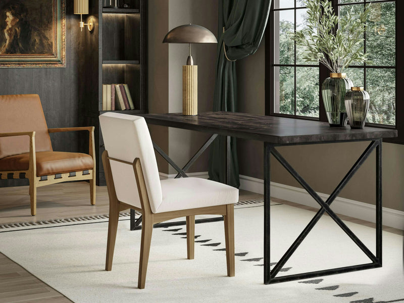 A modern home office featuring the Luca Desk in Charred Ember by James+James, paired with a white upholstered chair. A lamp and decorative vases adorn the desk, while a bookshelf with books and a framed painting stand to the left next to a brown leather chair. A large window lets in natural light.