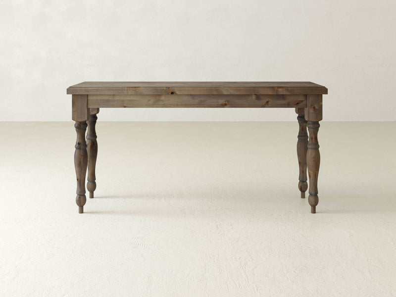 An Abigail Sofa Table - Barn Wood from James+James, featuring a rectangular top and four intricately carved legs, stands on a light-colored floor against a plain, light-colored wall. The table has a rustic finish that emphasizes the natural grain of the wood.