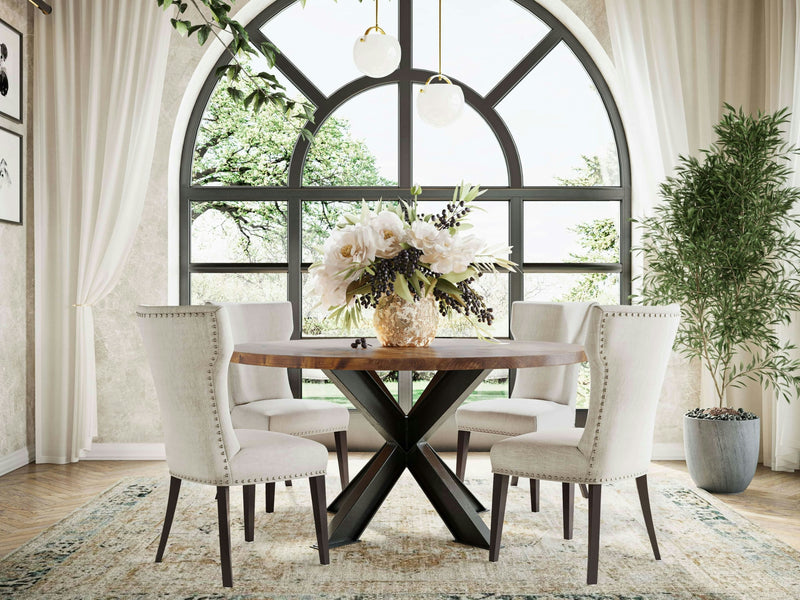 A dining room with the Shiloh Round Dining Table - Tuscany by James+James surrounded by four upholstered chairs. A vase of flowers sits on the table, which stands on a patterned rug. The room features a large arched window, sheer white curtains, potted plants, and ample natural light.