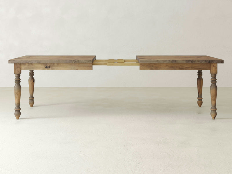 A French Country Expandable Dining Table in Harvest Wheat from James+James stands on a light-colored floor against a plain wall. The table features four turned legs and an extension mechanism in the middle with a visible wooden support system. The tabletop is split, showcasing the extended section.