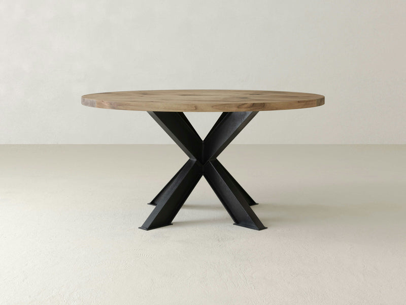 The Shiloh Round Dining Table - Harvest Wheat by James+James features a light beige wooden top and black metal legs that intersect in an 'X' shape. Set against a plain, light-colored wall and floor, this table exudes a minimalist and modern design.