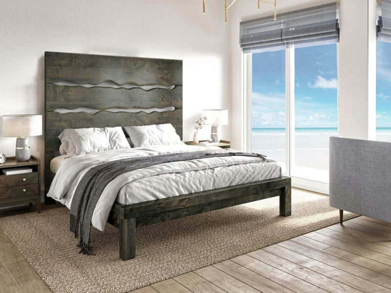 Modern bedroom with wooden furniture featuring the Live Edge Bed - Deep Grey by James+James, showcasing a dark wood frame and headboard. The room has large windows offering a stunning ocean view. A gray sofa sits near the bed, and the space is lit by contemporary lamps on bedside tables.