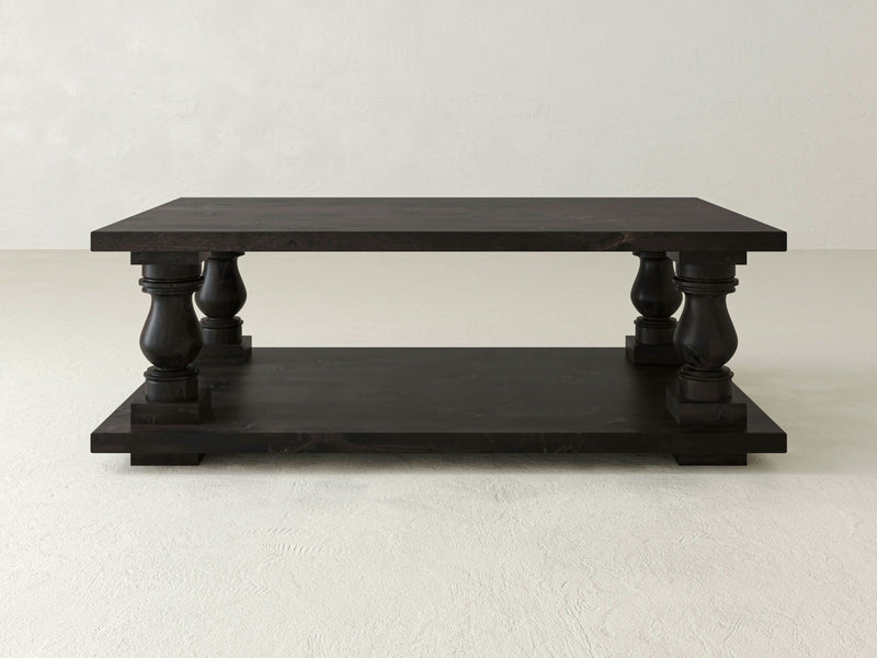 A Vivien Coffee Table - Charred Ember from James+James standing on a light-colored floor. The table has a rectangular top with paneled legs featuring ornate carvings. Below the top surface, there is a lower shelf providing additional storage space.