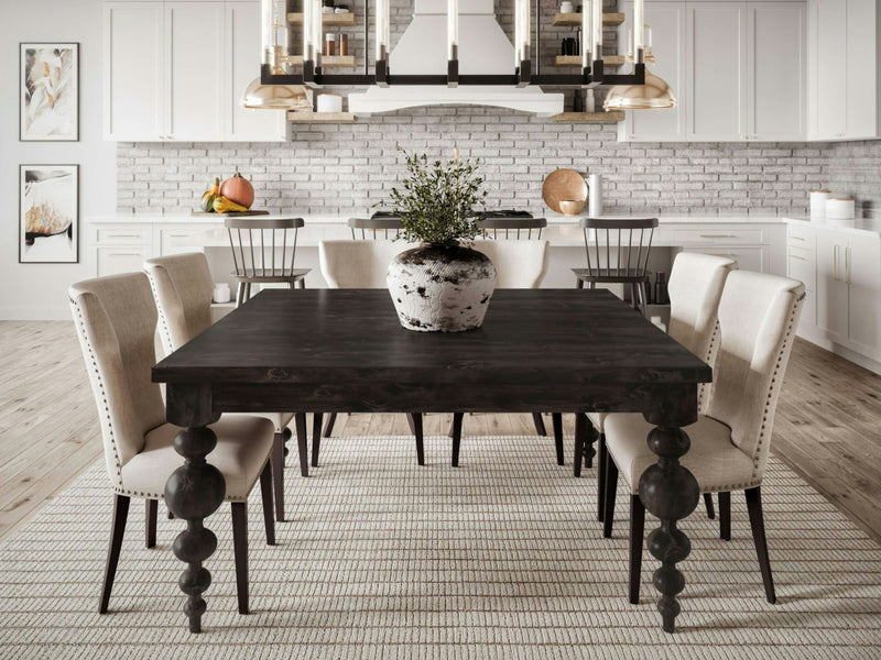 A modern dining room with an Olivia Square Dining Table in Charred Ember by James+James, surrounded by six cream-colored upholstered chairs. A large potted plant is centered on the table. The background features white cabinets, a light brick backsplash, and pendant lights. Two framed artworks hang on the wall.