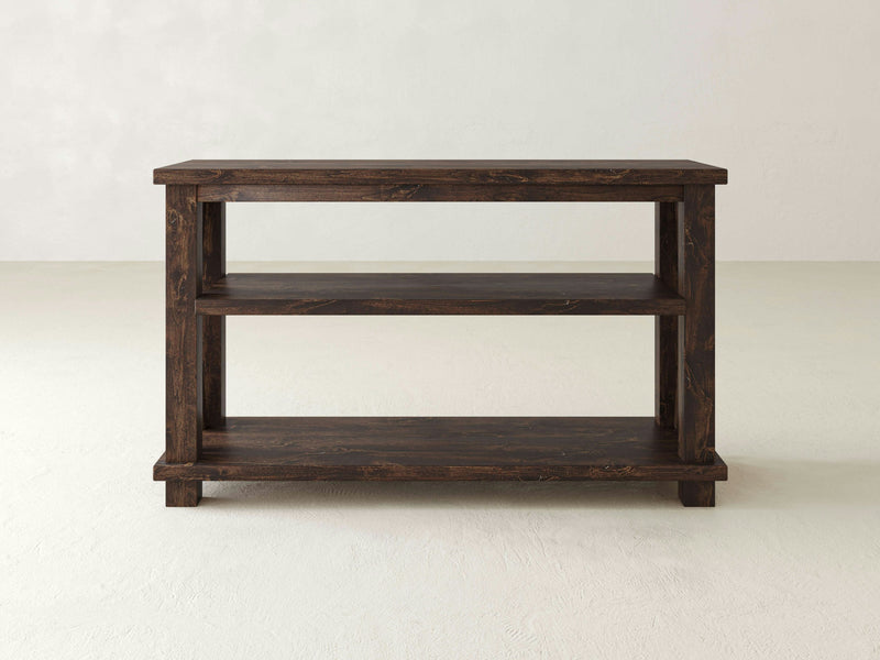 The Carter X Sofa Table by James+James features a rich tobacco finish. This wooden console table includes three shelves, including the top surface, and boasts an open design. Positioned against a light, neutral background, it showcases a simple and rustic style.