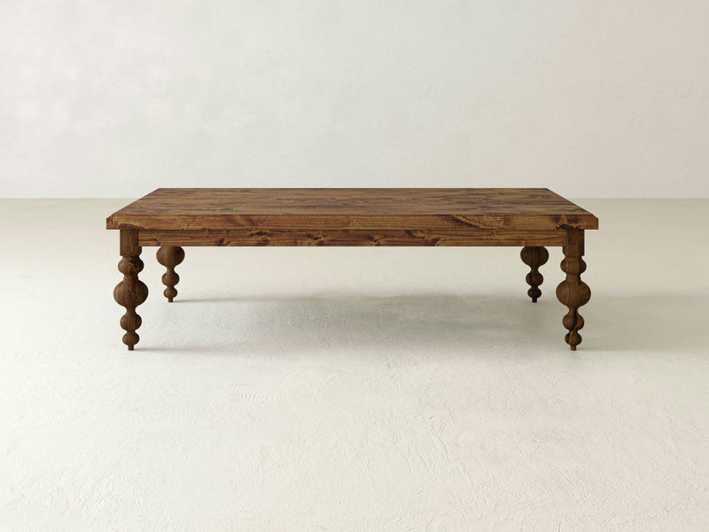 An Olivia Coffee Table - Tuscany from James+James, featuring a rustic wooden design with intricately carved legs, placed in an empty, minimalistic room with a white floor and light gray walls. The table's surface showcases a natural wood grain finish.