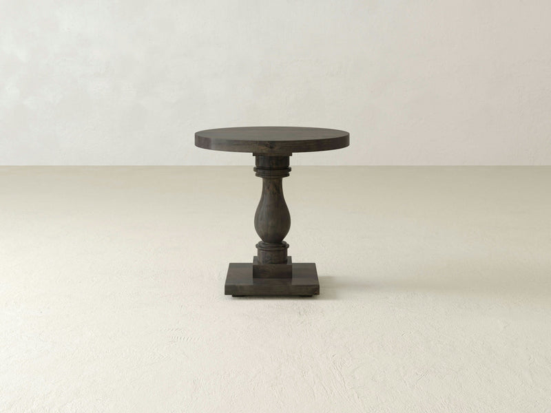 The Vivien Side Table - Deep Grey by James+James is a small, round wooden side table featuring a single central pedestal leg with a square base. It stands in an empty, light-colored room with a smooth floor and a plain wall background. The table boasts a simple and minimalist design.