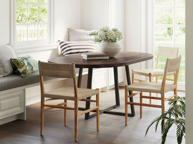 A bright dining room features the Watson Oval Dining Table - Tobacco by James+James, three beige chairs, and a built-in cushioned bench adorned with pillows. A large vase with white flowers graces the table, while large windows let in natural light, illuminating the space and highlighting a plant placed by the window.