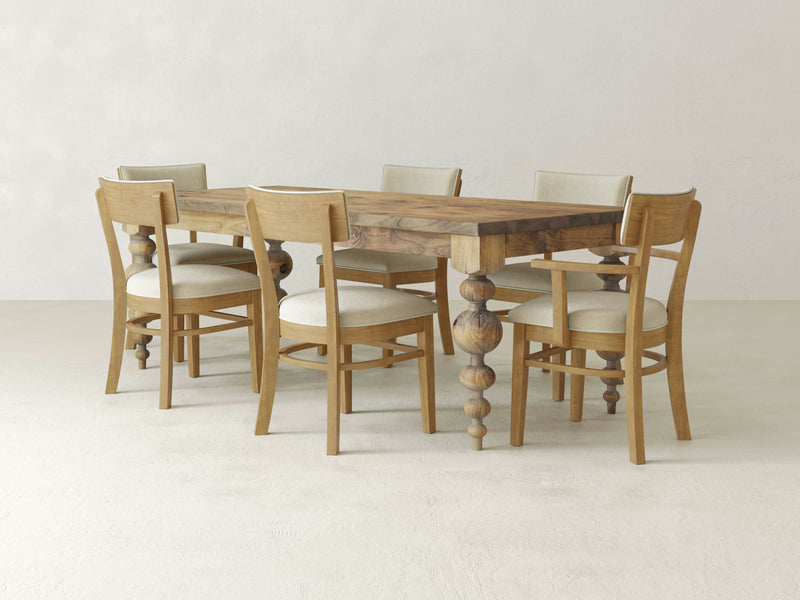 The Olivia & Everly Dining Set by James & James features the Olivia Dining Table with intricate, turned legs, accompanied by six matching Everly Dining Chairs. The chairs boast light-colored, cushioned seats and backrests that perfectly complement the table's natural wood finish. This elegant dining set is showcased in a minimalistic, neutral-toned room reminiscent of harvest wheat.