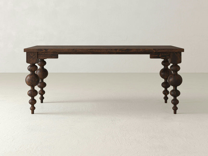 The Olivia Desk - Tobacco by James+James, featuring dark wooden construction and intricately carved legs with spherical and cylindrical shapes, is set against a plain, light-colored background. Its simple rectangular top combined with the detailed craftsmanship on the legs gives it an elegant antique appearance.