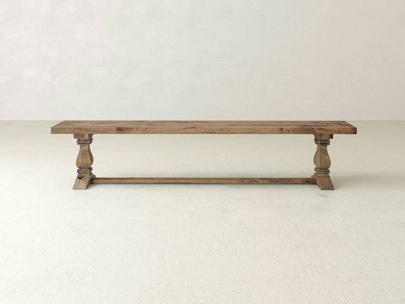 The Heirloom Bench - Harvest Wheat by James+James is centered against a light, plain background. This rustic wooden bench, with its simple and natural finish, features turned legs and a horizontal support bar near the base, highlighting its classic design.