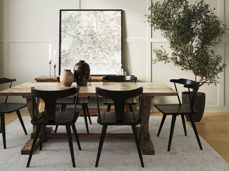 A modern dining room showcases the Vera Expandable Dining Table from James+James in barn wood, accompanied by six sleek black chairs. The back wall is adorned with a framed abstract artwork and two illuminated candlesticks. To the right, an indoor potted tree adds a touch of greenery to the space.