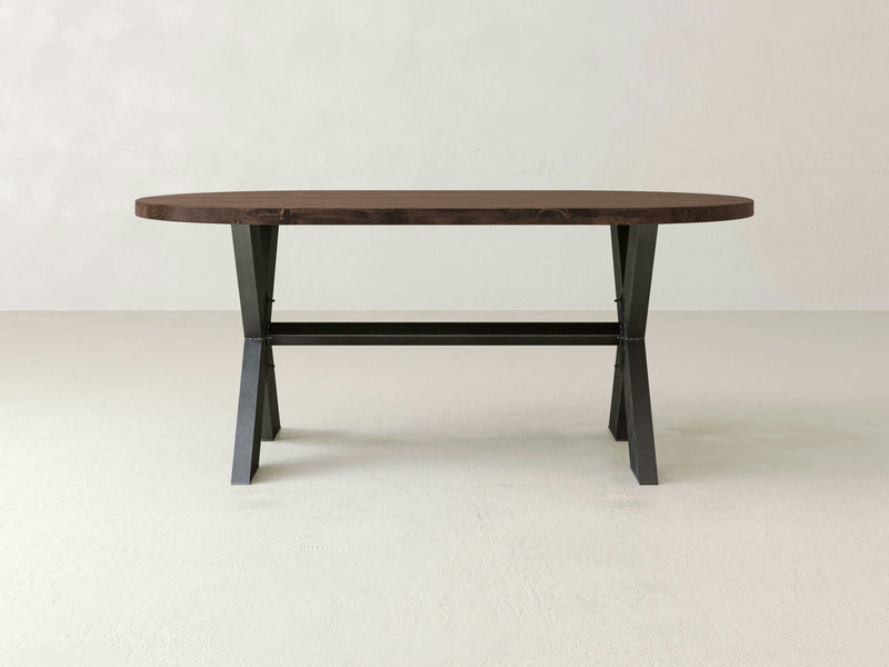 The X-Base Oval Dining Table - Tobacco from James+James features a dark brown wooden top and black metal legs, captured against a plain, light-colored background. The table boasts a simple, modern design with a T-shaped support structure connecting the legs.