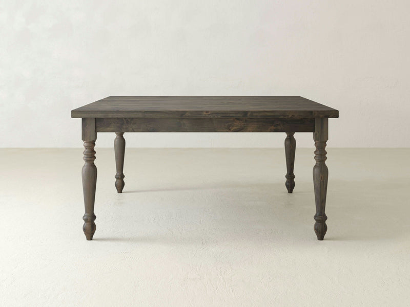 A French Country Square Dining Table in deep grey, crafted by James+James, is centered in a minimalistic room with a light-colored floor and off-white walls. The table features four ornate, turned legs and a plain rectangular top, showcasing a simple yet elegant design.