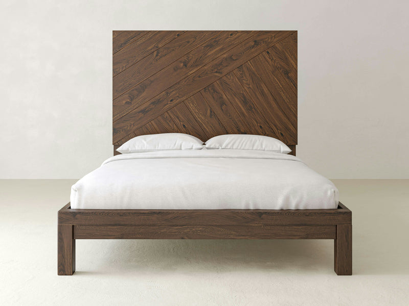 The Luna - Deep Grey bedframe by James+James is a minimalist piece featuring a tall, decorative headboard with a crisscross pattern. The bed is adorned with crisp white bedding, including two pillows. The plain, light-colored wall and floor in the background emphasize the bed's simple and elegant design.