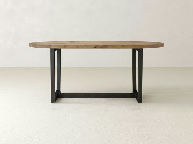 A sleek and modern Trapezoid Oval Dining Table - Harvest Wheat by James+James, featuring an oval-shaped wooden top with minimalist, dark-colored metal legs, is centered against a light-colored background.