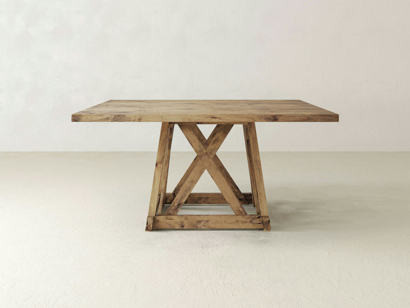 The Julia Square Dining Table from James+James features a sturdy, rectangular top and a distinct, geometric X-shaped base design. The table is placed on a light-colored floor against a plain, off-white wall. The wood has a rustic finish and showcases its natural grain, reflecting the Harvest Wheat color.