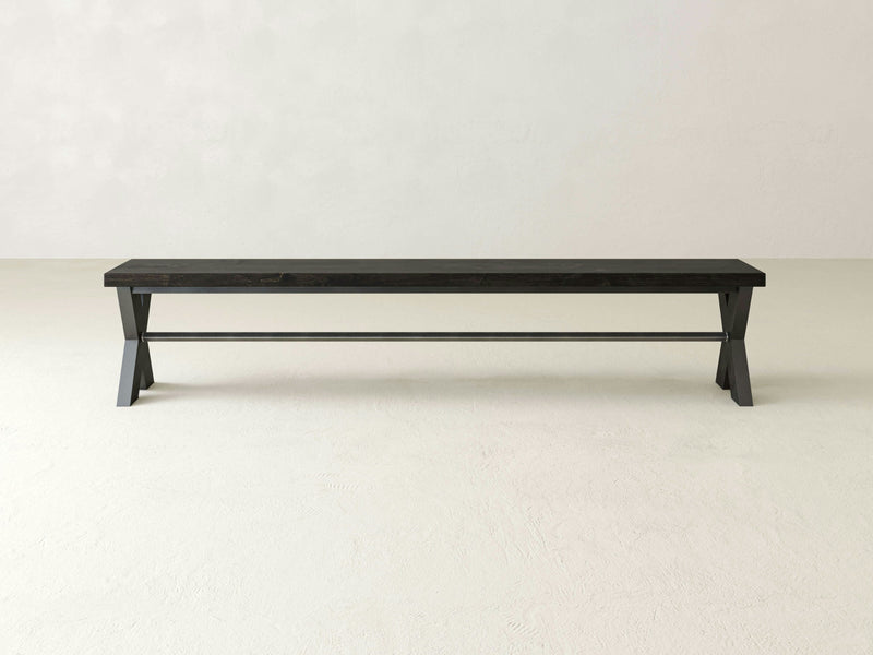 The X-Base Bench - Charred Ember by James+James stands on a light-colored, textured floor. The minimalist wooden bench features simple, straight lines and is supported by two sturdy legs, each forming a triangular shape with a connecting horizontal bar for added stability.