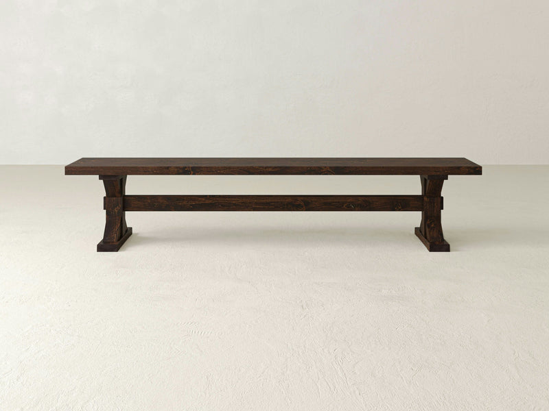 A James+James Trestle Bench, featuring a long, dark wooden structure with a simple, rectangular design, is placed on a light-textured floor against a plain, off-white wall. The bench boasts sturdy block-style legs connected by a horizontal support beam, enhancing the space with a touch of farmhouse style.