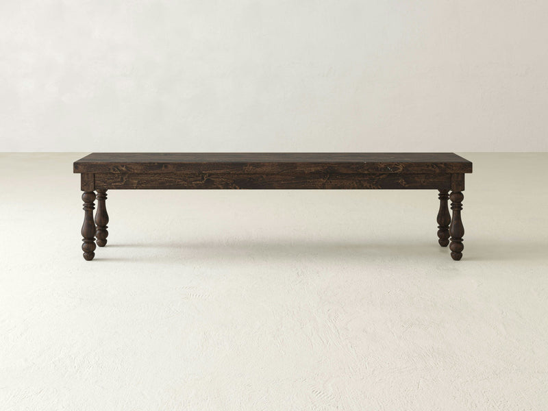 The Annli Bench - Tobacco by James+James, characterized by its rectangular top and intricately carved legs, is placed against a plain, off-white background. The bench boasts a rustic, classic design crafted from rich, dark-stained wood.