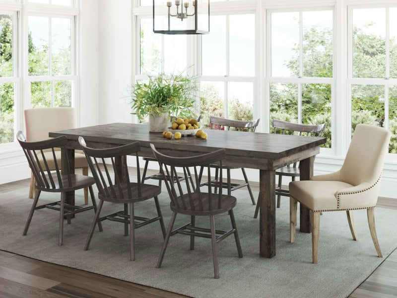 Bright dining area with large windows showcasing a lush green view. The James+James Farmhouse Expandable Dining Table in Deep Grey is surrounded by six chairs: two upholstered chairs with nailhead trim and four wooden chairs. A bowl of lemons and a plant centerpiece sit on the table under a contemporary light fixture.