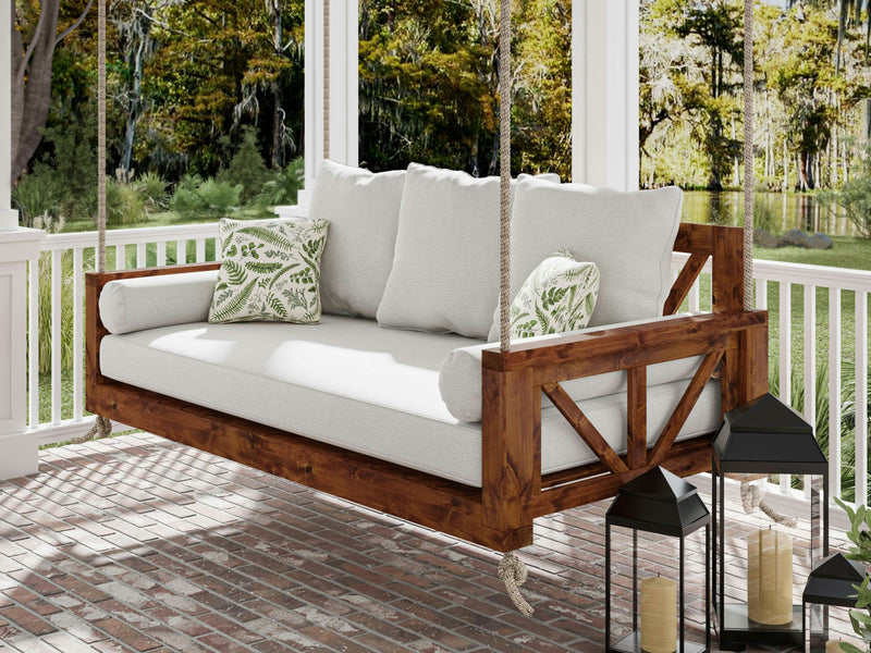 A close-up of the Avery Porch Swing - Tuscany from James+James features a wooden seat with a white cushion on top. The swing is secured by a thick rope threaded through a hole in the wooden armrest, and you can see a hint of a striped rug on the floor beneath.