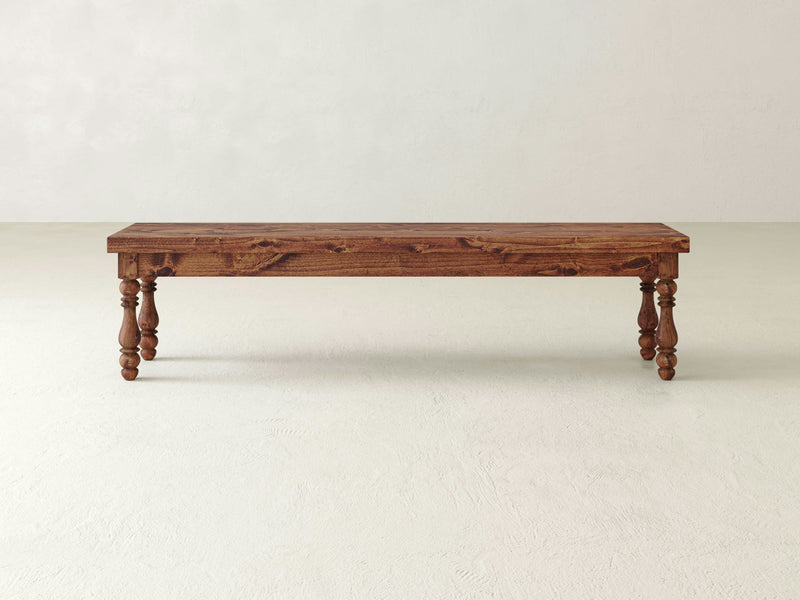 A long, rectangular wooden Annli Bench - Tuscany from James+James with a polished surface and intricately carved legs. The bench is set against a plain, light-colored background on a smooth floor.