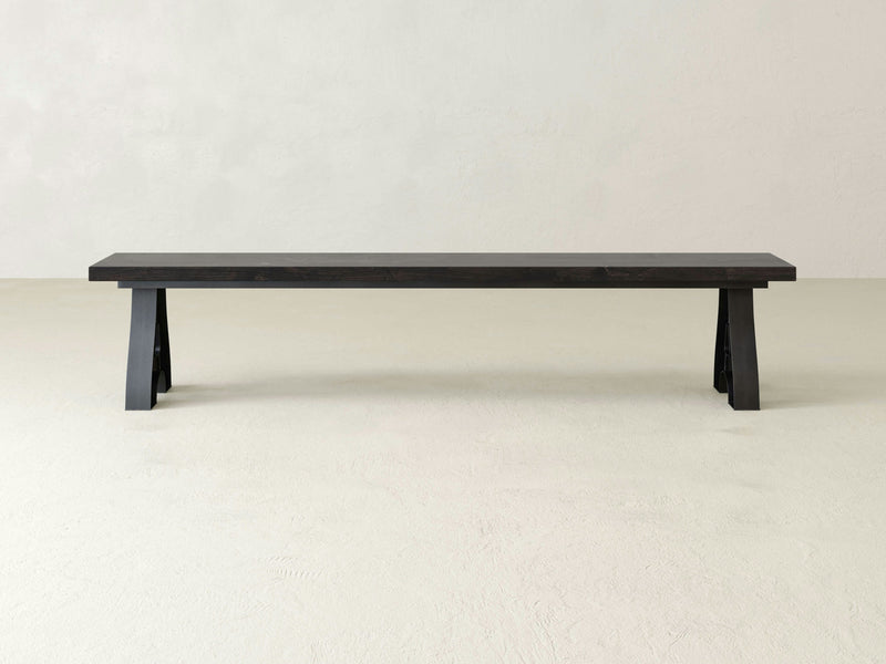 The Wishbone Bench - Charred Ember by James+James, featuring sturdy rectangular legs and a smooth, flat seat surface, is placed against a light-colored background.