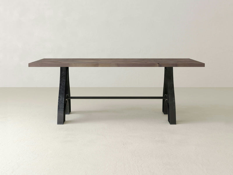 The Wishbone Dining Table by James+James exudes modern dining room elegance with its vintage industrial minimalist design, featuring a rectangular wooden top and black metal A-shaped legs. It stands on a light gray floor, set against an off-white wall.