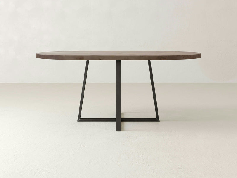 The Watson Oval Dining Table - Barn Wood by James+James showcases a minimalist design with its dark brown wooden top and black metal base that features a central support and angled legs. It stands out beautifully on a light-colored floor against a neutral wall background, embodying simplicity and modernity.