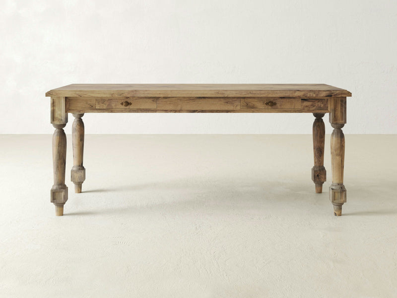 The Rosemary Desk - Harvest Wheat from James+James is a rustic wooden table featuring a rectangular top and four intricately carved legs. The wood boasts a weathered, natural finish, exuding a farmhouse-style charm. The desk is set against a plain white background.