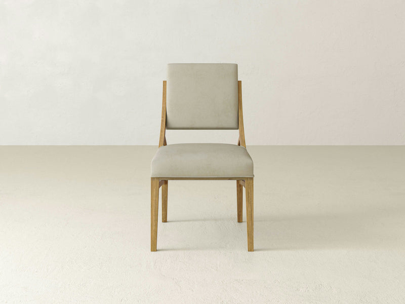 The Quinn Dining Chair - Harvest Wheat by James+James is centered in a minimalist setting, showcasing its light beige upholstered seat and backrest. The background features a neutral, lightly textured wall and a clean, light-colored floor.