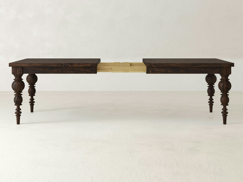 The Bailey Expandable Dining Table by James+James is depicted partially extended, showcasing its intricate, modern turned legs against a dark wooden finish. The center section of the table is open, revealing a wooden extension mechanism designed for lengthening the table. A plain, light-colored wall and floor form the backdrop, ideal for a sophisticated dining space.