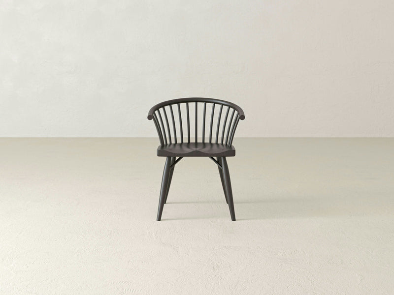 The James+James Maria Dining Chair, made from solid wood, boasts a sleek black finish with a curved backrest and slender vertical supports. It stands on four tapered legs in an empty, light-colored room. Its contoured seat and minimalist design perfectly complement the plain background wall adorned with subtle texture.